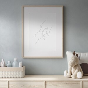 Puede incluir: Impresi&oacute;n enmarcada de arte lineal que representa la mano de un padre sosteniendo la mano de un beb&eacute;. La obra de arte est&aacute; en un marco de madera clara y cuelga de una pared azul claro. Un peluche y objetos decorativos est&aacute;n sobre una superficie de madera debajo.