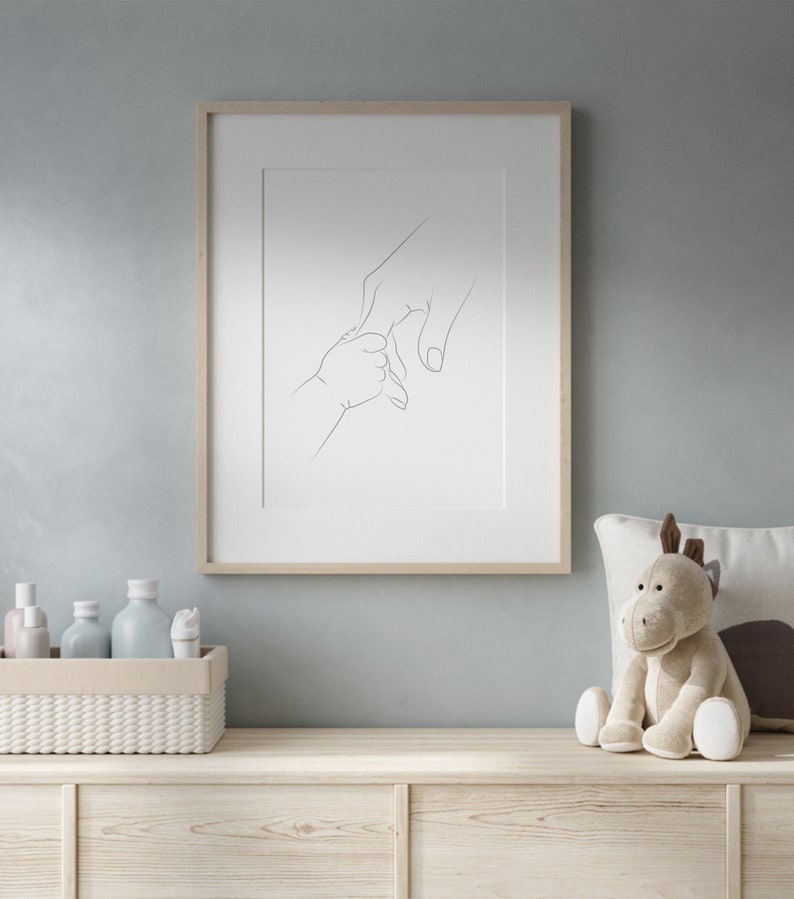 Puede incluir: Impresi&oacute;n enmarcada de arte lineal que representa la mano de un padre sosteniendo la mano de un beb&eacute;. La obra de arte est&aacute; en un marco de madera clara y cuelga de una pared azul claro. Un peluche y objetos decorativos est&aacute;n sobre una superficie de madera debajo.