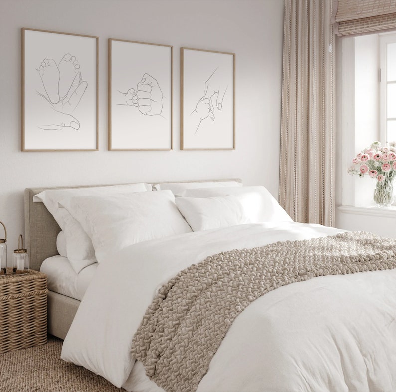 Puede incluir: Tres impresiones de arte de l&iacute;nea minimalista en marcos de madera que cuelgan de una pared blanca sobre una cama blanca con una manta de tiro beige. Las impresiones representan una mano sosteniendo el pie de un beb&eacute;, una mano sosteniendo el pu&ntilde;o de un beb&eacute; y una mano sosteniendo el dedo de un beb&eacute;.