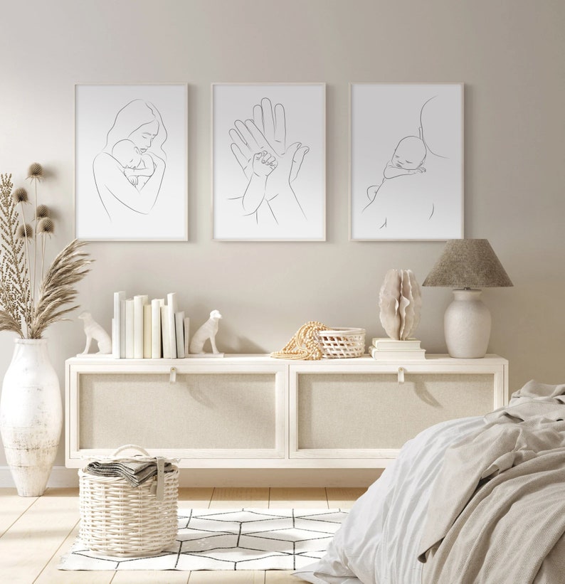 Puede incluir: Tres impresiones minimalistas de arte lineal en marcos blancos que representan a una madre sosteniendo un beb&eacute;, dos manos sosteniendo la mano de un beb&eacute; y un padre llevando a un beb&eacute; dormido sobre su hombro. Las impresiones se muestran en una pared blanca sobre una c&oacute;moda blanca con una l&aacute;mpara blanca en el lado derecho.