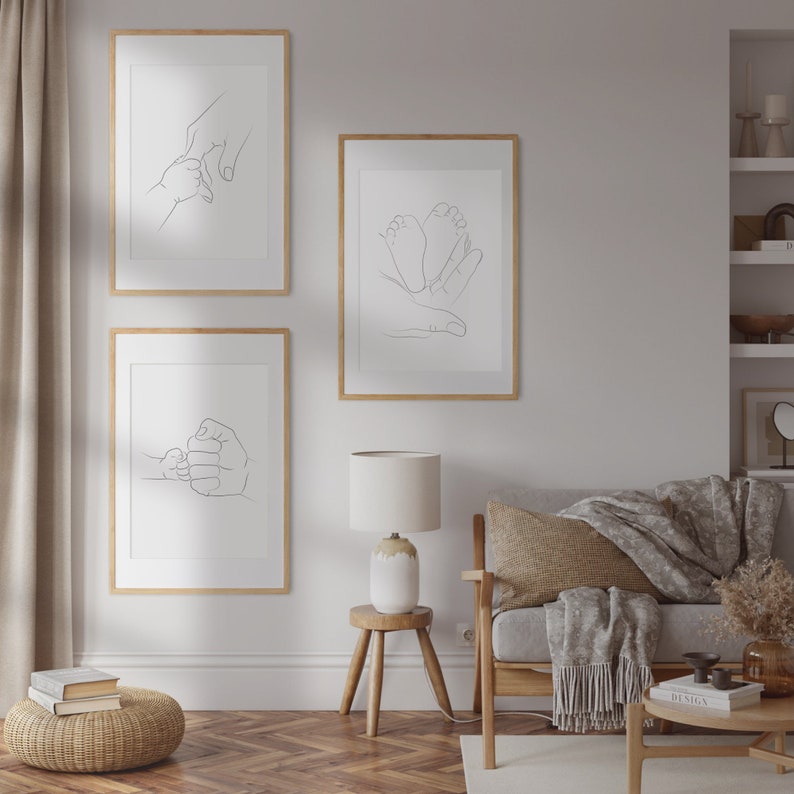 Puede incluir: Tres impresiones de arte de l&iacute;nea minimalista en marcos de madera, que representan a un padre y un hijo d&aacute;ndose la mano, un padre y un hijo d&aacute;ndose el pu&ntilde;o y un padre sosteniendo los pies de un beb&eacute;. Las impresiones se muestran en una pared blanca sobre un sof&aacute; de madera con una manta y una mesa auxiliar de madera con una l&aacute;mpara.