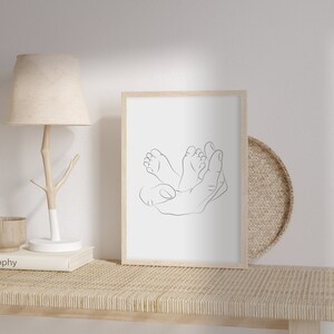 Puede incluir: Una impresi&oacute;n enmarcada de arte lineal que presenta una ilustraci&oacute;n minimalista de una mano que sostiene dos pies peque&ntilde;os. La obra de arte est&aacute; en un marco de madera clara y descansa sobre una estanter&iacute;a tejida, junto a una l&aacute;mpara y objetos decorativos. La est&eacute;tica general es neutra y relajante.