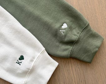 Puede incluir: Dos sudaderas con corazones y letras bordadas. Una es blanca con un corazón verde y la letra K. La otra es verde con un corazón blanco y la letra A.