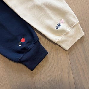 May include: Close-up of two sweatshirt sleeves. One is navy blue with a small embroidered "e" and a red heart. The other is cream-colored with an embroidered "M" and a pink heart. The cuffs are ribbed.