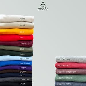 May include: A stack of folded t-shirts in various colors, including white, sand, gold, red, burgundy, olive, forest, light blue, royal blue, purple, brown, navy, charcoal, and black. The text "PINE GOODS" is visible at the top of the image.