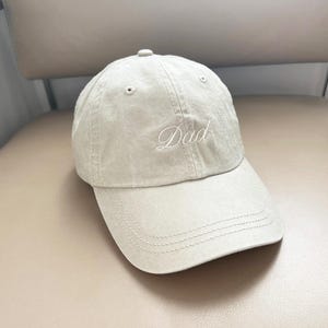 May include: A beige baseball cap with a white embroidered "Dad" on the front.