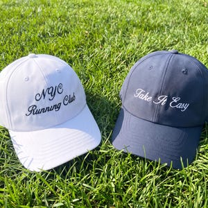May include: A white baseball cap with the black embroidered text NYC Running Club and a navy blue baseball cap with the white embroidered text Take It Easy. Both caps feature a structured crown, a curved brim, and a button on the top center.