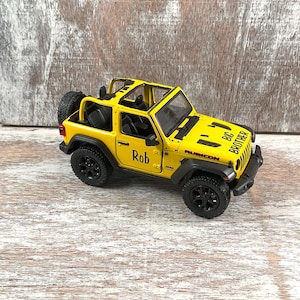 May include: A yellow toy Jeep Wrangler Rubicon with black wheels and a spare tire. The text "Rob" is on the side, and "BIG BROTHER" is on the hood. The toy is on a wooden surface.
