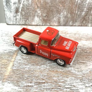 May include: A red toy pickup truck with silver wheels and a tan bed. The truck has the word "Poppy" on the door and "Ring Bearer" on the hood. Two gold wedding rings are on the roof.