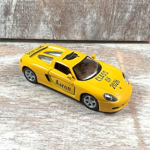 Puede incluir: Un coche de juguete amarillo con el texto "CLASS OF 2026" y "Aaron" impreso. El coche tiene un gorro de graduaci&oacute;n negro encima. El coche est&aacute; sobre una superficie de madera blanca y gris.
