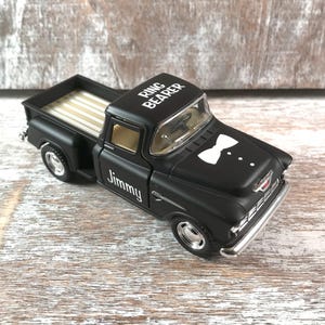 May include: A black toy pickup truck with the words "Ring Bearer" and "Jimmy" printed on it. The truck has a white bow tie graphic on the front and a tan bed. The truck is on a wooden surface.