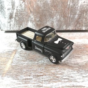 May include: A black toy truck with the words "Ring Bearer" on the roof and "Jimmy" on the side. The truck has a white bow tie design on the front and a striped bed.