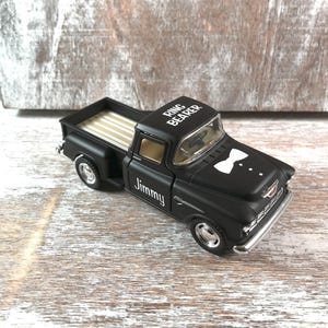 May include: A black toy pickup truck with the words "Ring Bearer" on the roof and "Jimmy" on the side. The truck has a white bow tie graphic on the front and a tan bed.
