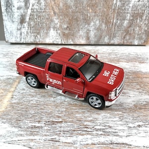 May include: A red toy pickup truck with the words "BIG BROTHER" and "Payson" printed on it. The truck has a sunroof, chrome accents, and silver wheels. The truck is sitting on a light-colored wooden surface.