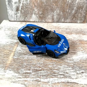 May include: A blue toy convertible car with the door open, displaying the name "HENRY". The car has black wheels and the text "RING BEARER" on the front. The car is on a wooden surface.