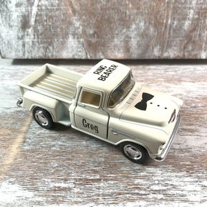 May include: A white toy truck with black wheels and chrome accents. The truck has 'Ring Bearer' printed on the roof and a black bow tie graphic on the hood. The name 'Greg' is printed on the door. The truck is sitting on a wooden surface.