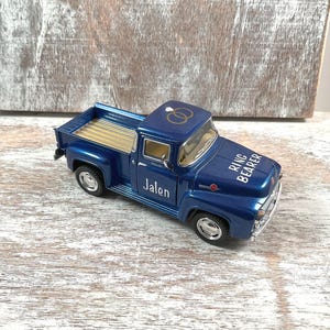 May include: A blue toy pickup truck with gold detailing in the bed. The truck has the name "Jalen" on the side and "Ring Bearer" printed on the hood. Two gold wedding rings are on the roof.