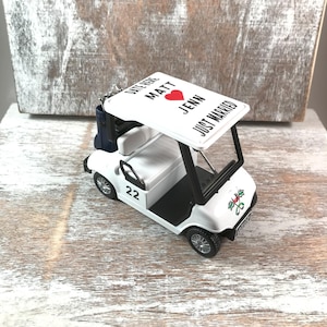 May include: A white golf cart wedding ornament with black accents. The cart features the number 22 on the side and a canopy with the words 'Date Here Matt Jenn Just Married' and a red heart. A small blue bag is attached to the back of the cart.