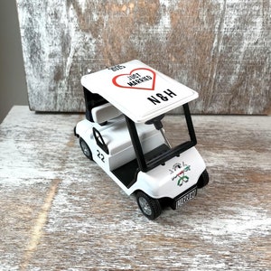 May include: White golf cart toy with a red heart that says "Just Married" and the initials "N&H" on the roof. The cart has a black steering wheel, black tires, and a black bumper. The cart has a number "22" on the side and the word "Married" on the back.