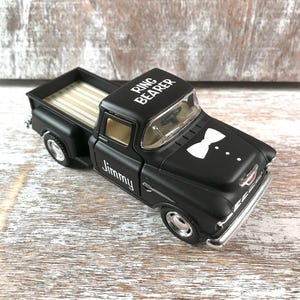 May include: A black toy truck with the words 'Ring Bearer' on the roof and 'Jimmy' on the side. The truck has a white bow tie design on the front. The truck is sitting on a light-colored wooden surface. The truck bed has a light-colored striped design.