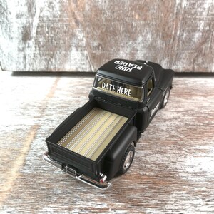 May include: A black toy pickup truck with a tan bed. The text "DATE HERE" is on the windshield and "RING BEARER" is on the roof. The truck has chrome accents and is set against a rustic wooden background.