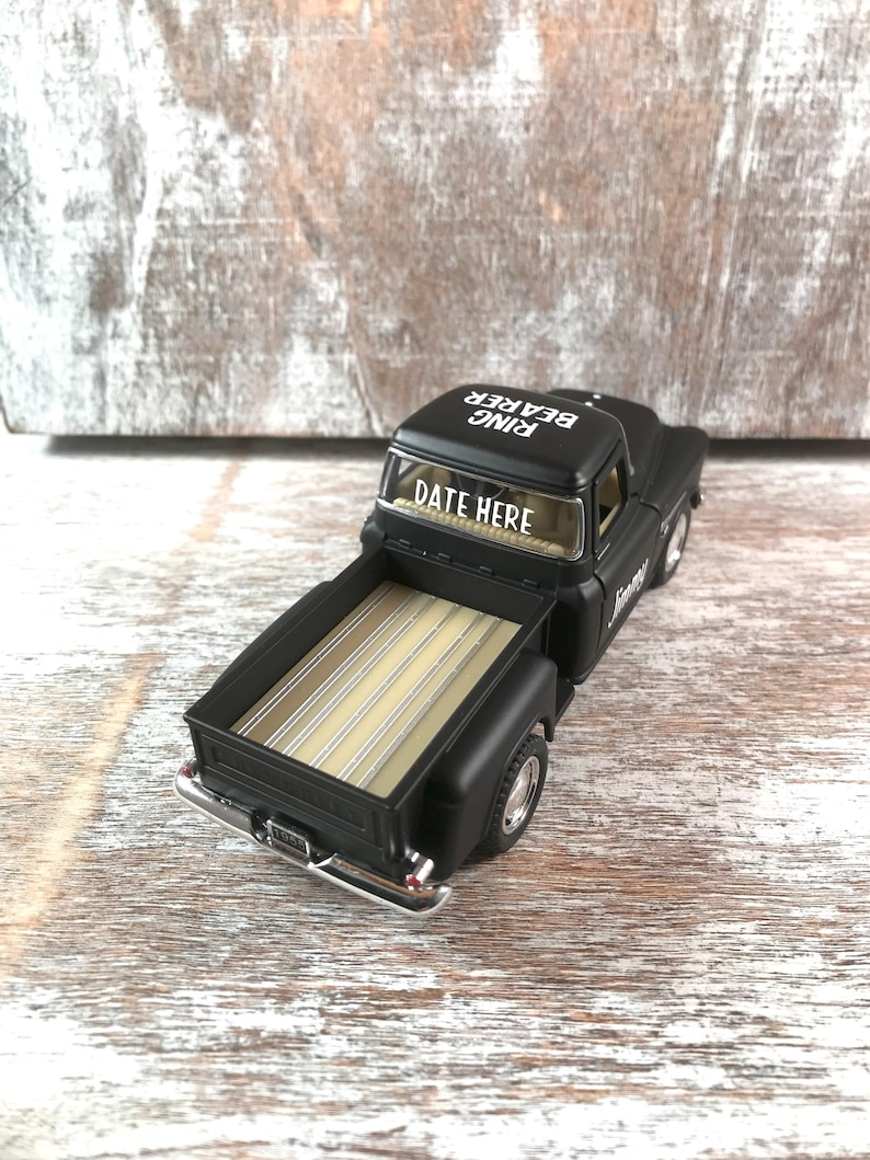 May include: A black toy pickup truck with a tan bed. The text "DATE HERE" is on the windshield and "RING BEARER" is on the roof. The truck has chrome accents and is set against a rustic wooden background.