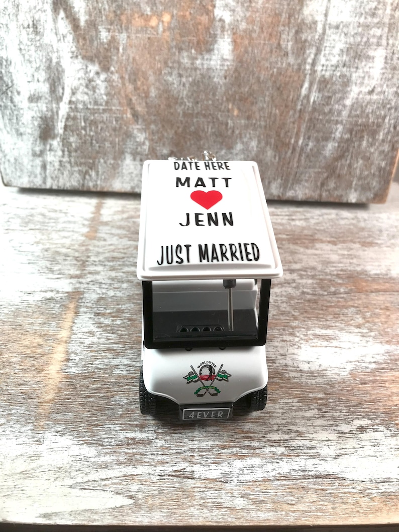 May include: A white golf cart ornament with the words "DATE HERE", "MATT", a red heart, "JENN", and "JUST MARRIED" printed on the roof. The front of the cart has the word "4EVER" on the license plate.