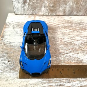 May include: A bright blue toy car with a black interior and a sleek design. The car is positioned on a wooden surface, with a ruler next to it. The car's length is approximately 3 inches.
