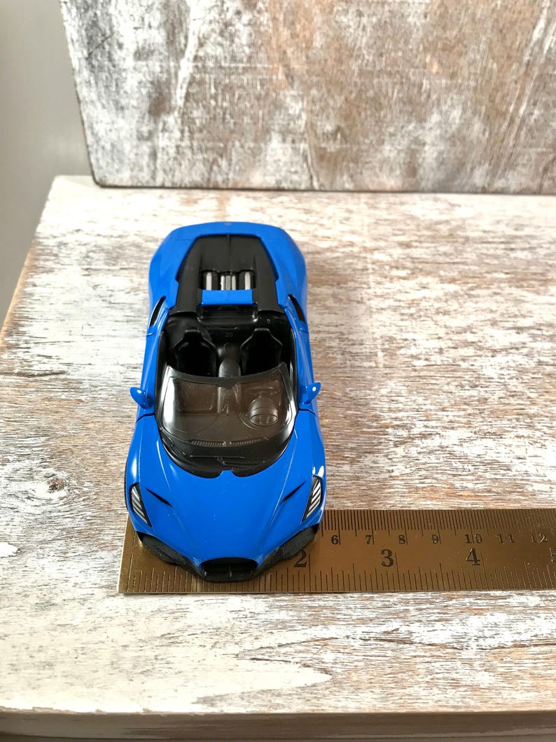 May include: A bright blue toy car with a black interior and a sleek design. The car is positioned on a wooden surface, with a ruler next to it. The car's length is approximately 3 inches.