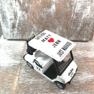 May include: A white golf cart ornament with black trim and wheels. The roof displays the text "DATE HERE MATT JENN JUST MARRIED" with a red heart. The number "22" is on the side. A festive design is on the front.