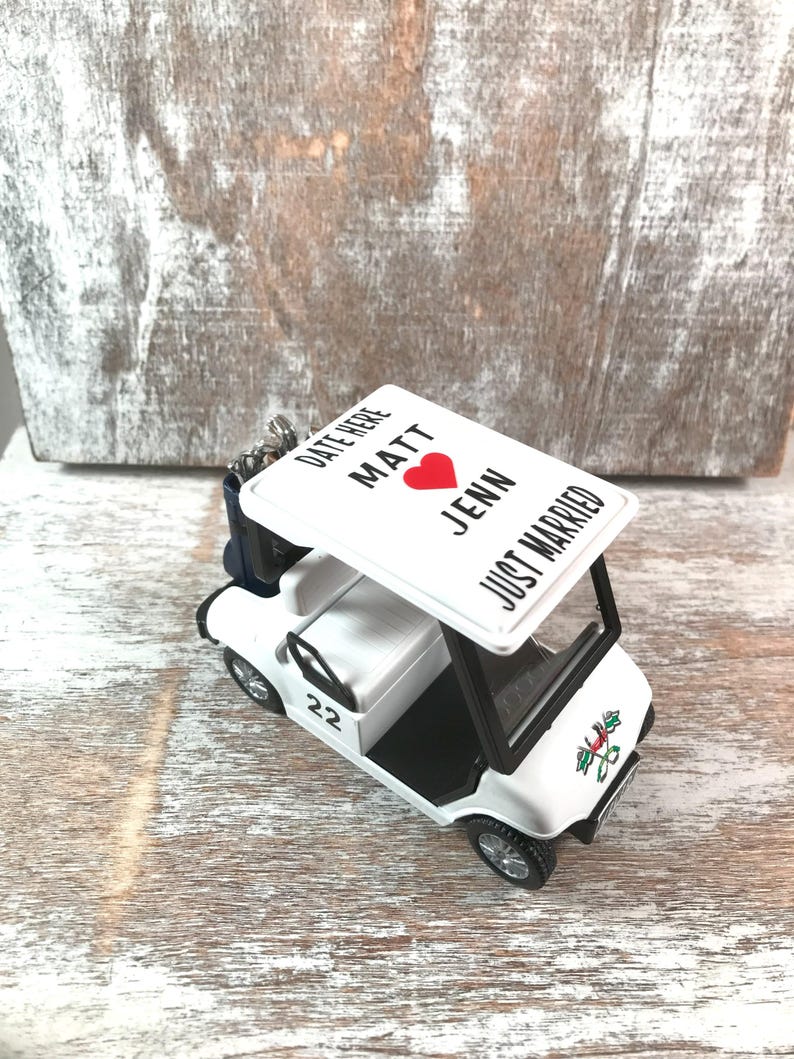 May include: A white golf cart ornament with black trim and wheels. The roof displays the text "DATE HERE MATT JENN JUST MARRIED" with a red heart. The number "22" is on the side. A festive design is on the front.