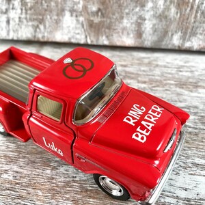 May include: A bright red toy pickup truck with the words "Ring Bearer" in white on the hood. The truck has a diamond ring graphic and interlocking rings on the roof. The name "Luke" is printed on the side.