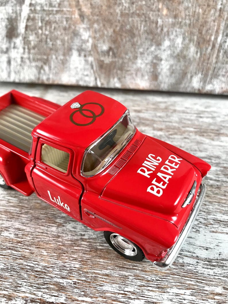 May include: A bright red toy pickup truck with the words "Ring Bearer" in white on the hood. The truck has a diamond ring graphic and interlocking rings on the roof. The name "Luke" is printed on the side.