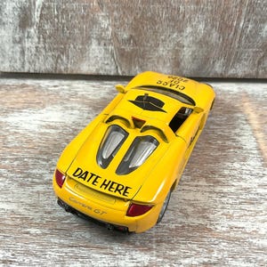 Puede incluir: Un coche de juguete amarillo brillante con el texto "DATE HERE" y "Carrera GT" en la parte trasera. El coche tambi&eacute;n tiene el texto "CLASS OF 2026" en la parte superior. El coche tiene detalles en negro y luces traseras rojas.