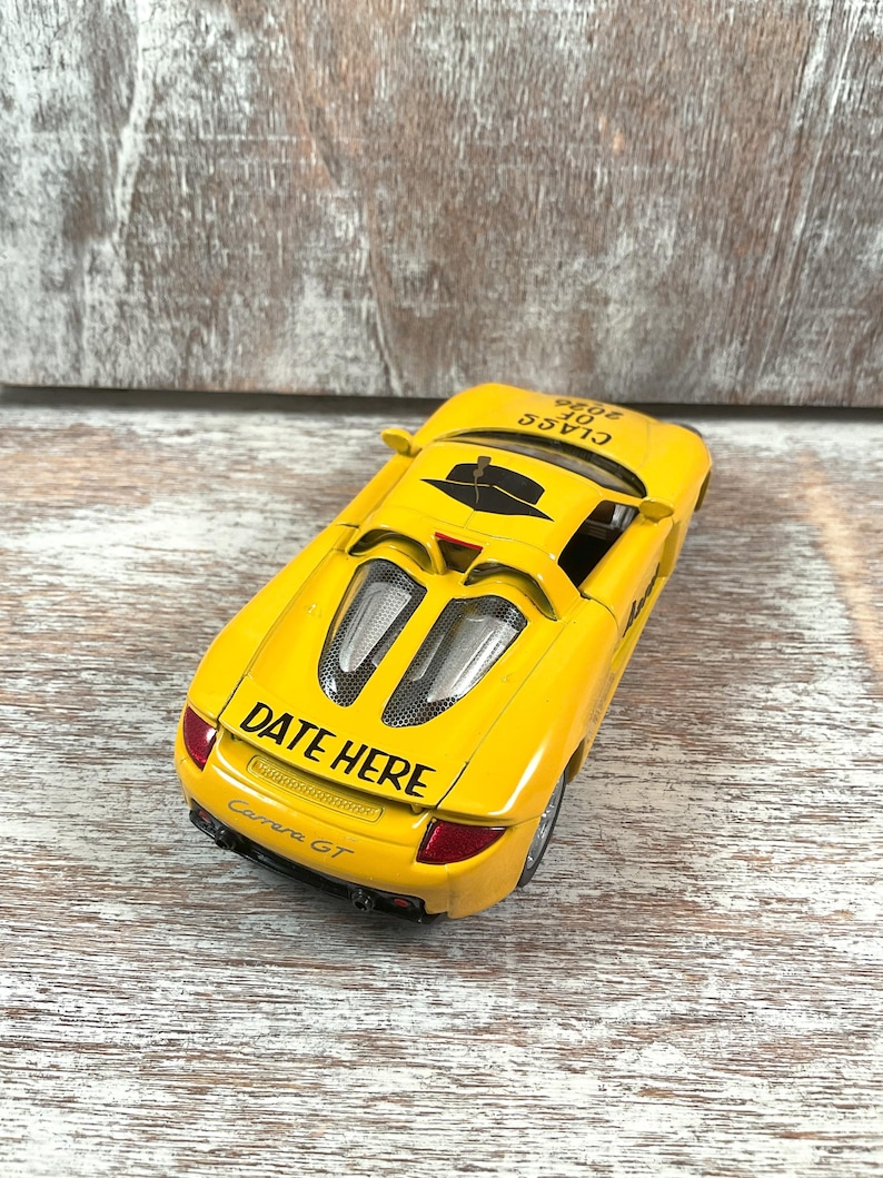 Puede incluir: Un coche de juguete amarillo brillante con el texto "DATE HERE" y "Carrera GT" en la parte trasera. El coche tambi&eacute;n tiene el texto "CLASS OF 2026" en la parte superior. El coche tiene detalles en negro y luces traseras rojas.
