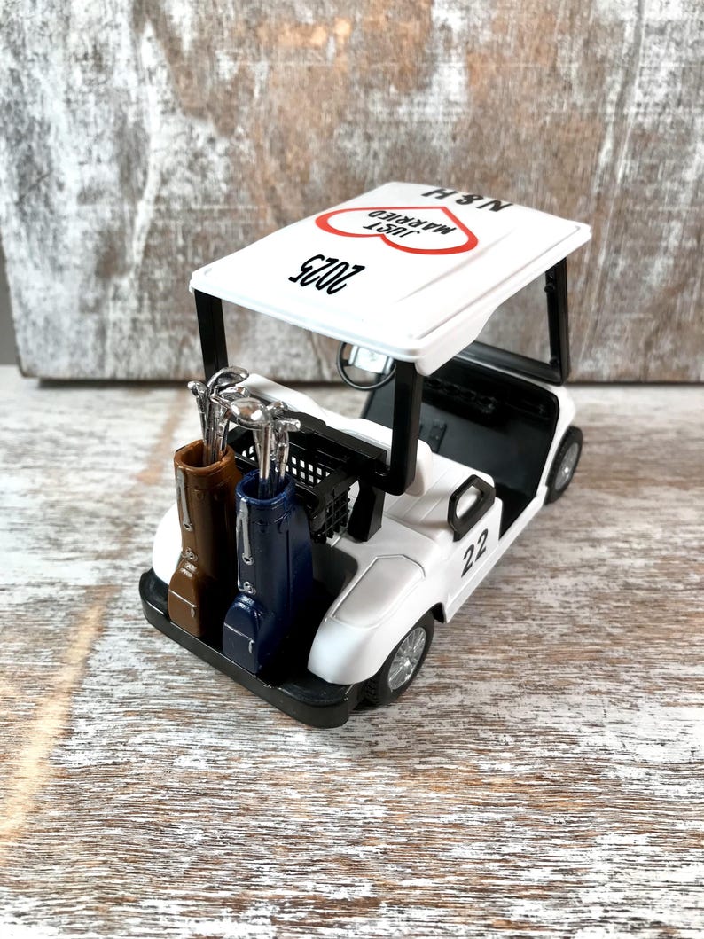 May include: White golf cart with a red heart and the text "Just Married" and "2023" on the roof. The cart has two golf bags and a number "22" on the side.