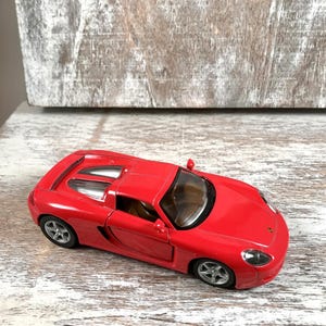 Puede incluir: Un coche deportivo de juguete rojo brillante con ruedas plateadas. El coche tiene un dise&ntilde;o elegante con un interior visible y un peque&ntilde;o aler&oacute;n. Est&aacute; sobre una superficie de madera blanca y gris.