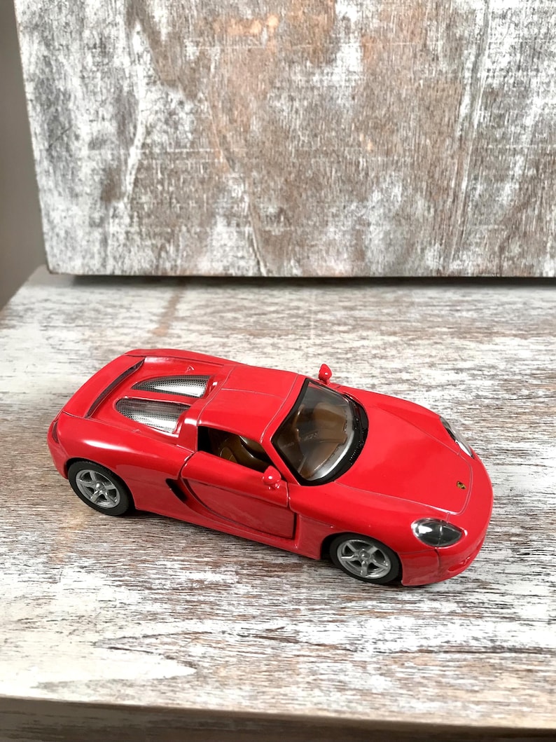 Puede incluir: Un coche deportivo de juguete rojo brillante con ruedas plateadas. El coche tiene un dise&ntilde;o elegante con un interior visible y un peque&ntilde;o aler&oacute;n. Est&aacute; sobre una superficie de madera blanca y gris.