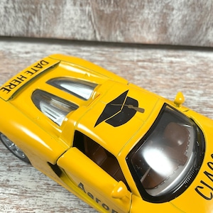 Puede incluir: Un coche de juguete amarillo con el texto "DATE HERE" en la parte trasera y "CLASS" en el lateral. El coche tiene un gr&aacute;fico de birrete negro en el techo y el nombre "Aaron" en el lateral.