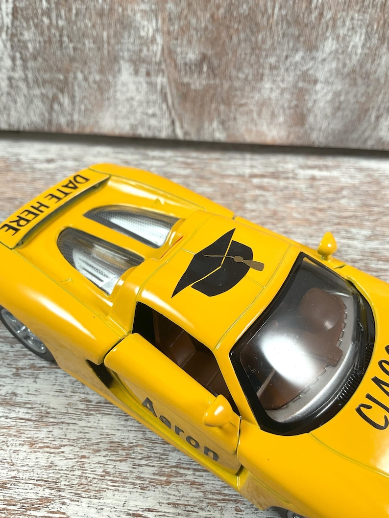 Puede incluir: Un coche de juguete amarillo con el texto "DATE HERE" en la parte trasera y "CLASS" en el lateral. El coche tiene un gr&aacute;fico de birrete negro en el techo y el nombre "Aaron" en el lateral.