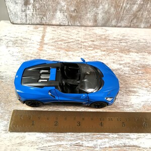 May include: A bright blue toy convertible car with black wheels and interior. The car is approximately 5 inches long and sits on a wooden surface. The car's design includes headlights and a detailed engine compartment.