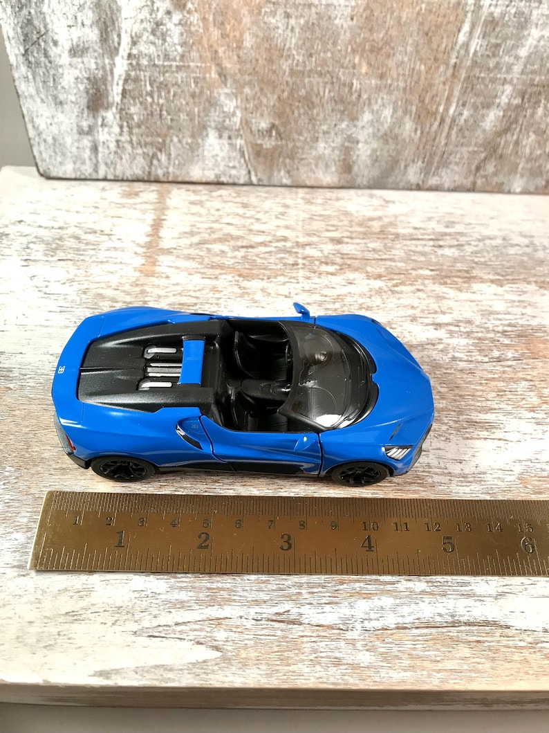 May include: A bright blue toy convertible car with black wheels and interior. The car is approximately 5 inches long and sits on a wooden surface. The car's design includes headlights and a detailed engine compartment.