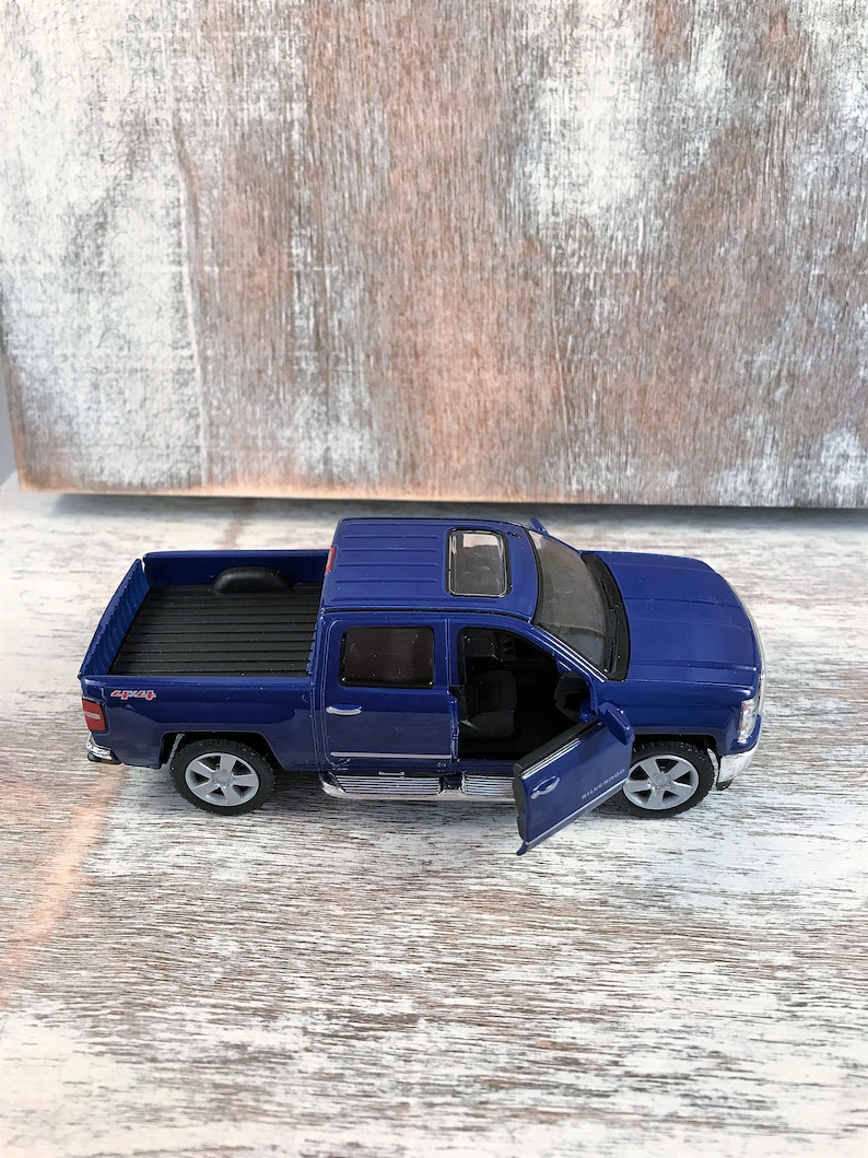 Może przedstawiać: Niebieska zabawka pick-up z otwartymi drzwiami od strony kierowcy. Samoch&oacute;d ma czarną skrzynię ładunkową i srebrne wykończenia. Koła są srebrne z sześcioramiennym wzorem. Samoch&oacute;d stoi na białej i szarej powierzchni.