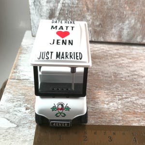 May include: A white miniature golf cart ornament with black accents. The top features the text "DATE HERE MATT JENN JUST MARRIED" with a red heart. The front reads "4EVER" and has a golf-themed emblem. The cart is approximately 4 inches long.