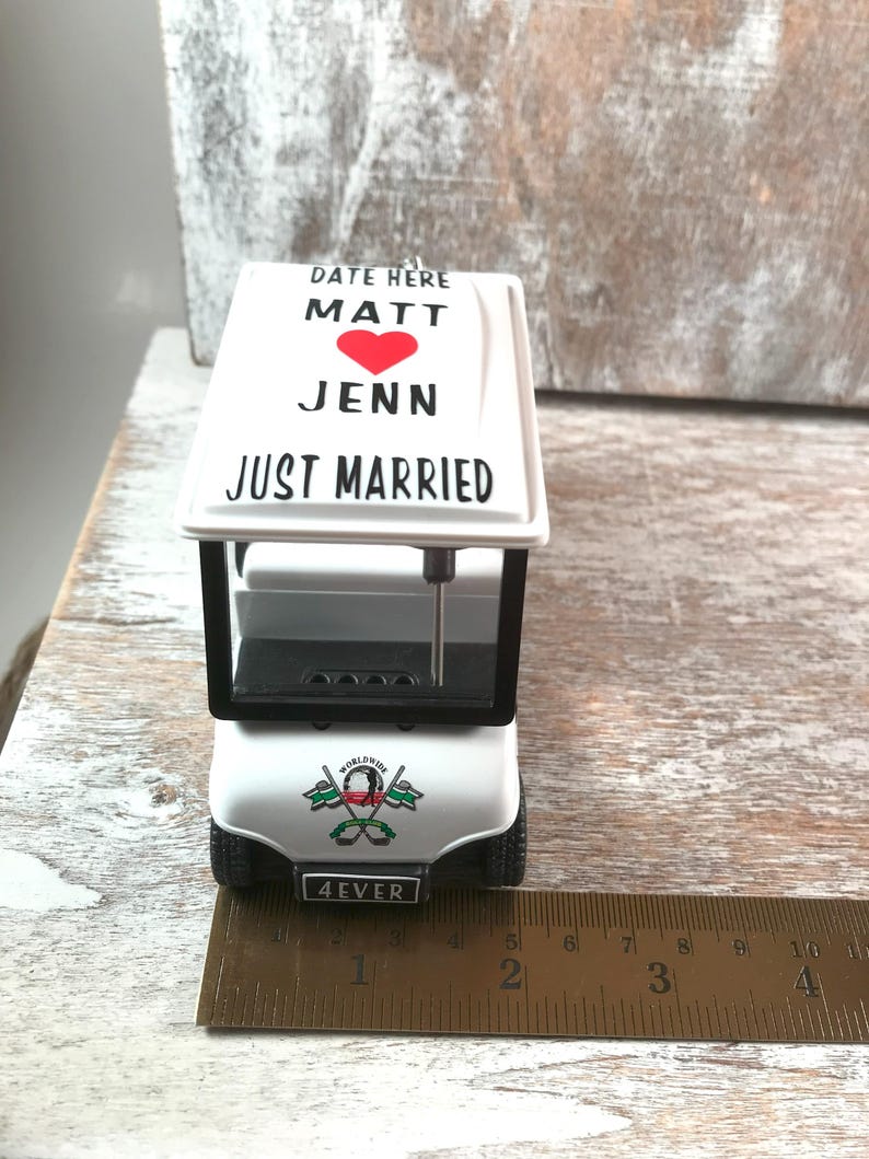 May include: A white miniature golf cart ornament with black accents. The top features the text "DATE HERE MATT JENN JUST MARRIED" with a red heart. The front reads "4EVER" and has a golf-themed emblem. The cart is approximately 4 inches long.