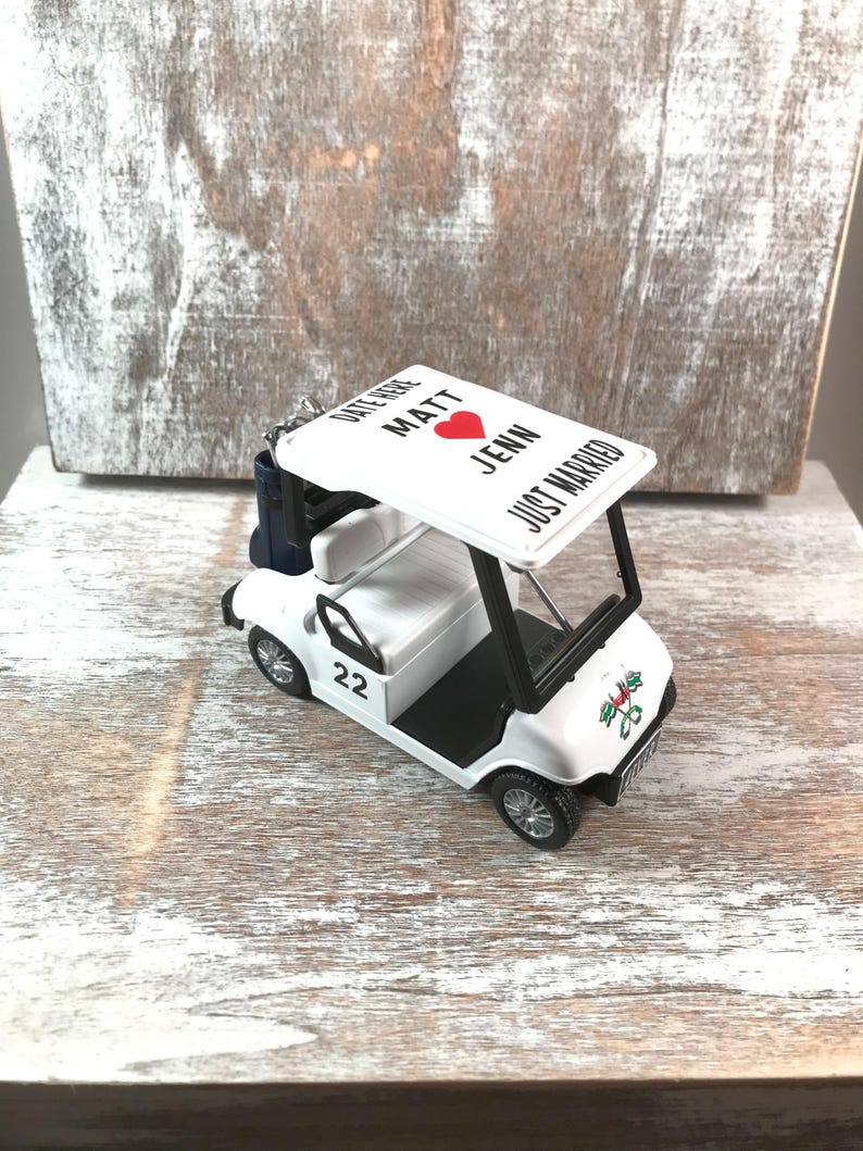May include: A white golf cart wedding ornament with black accents. The cart features the number 22 on the side and a canopy with the words 'Date Here Matt Jenn Just Married' and a red heart. A small blue bag is attached to the back of the cart.