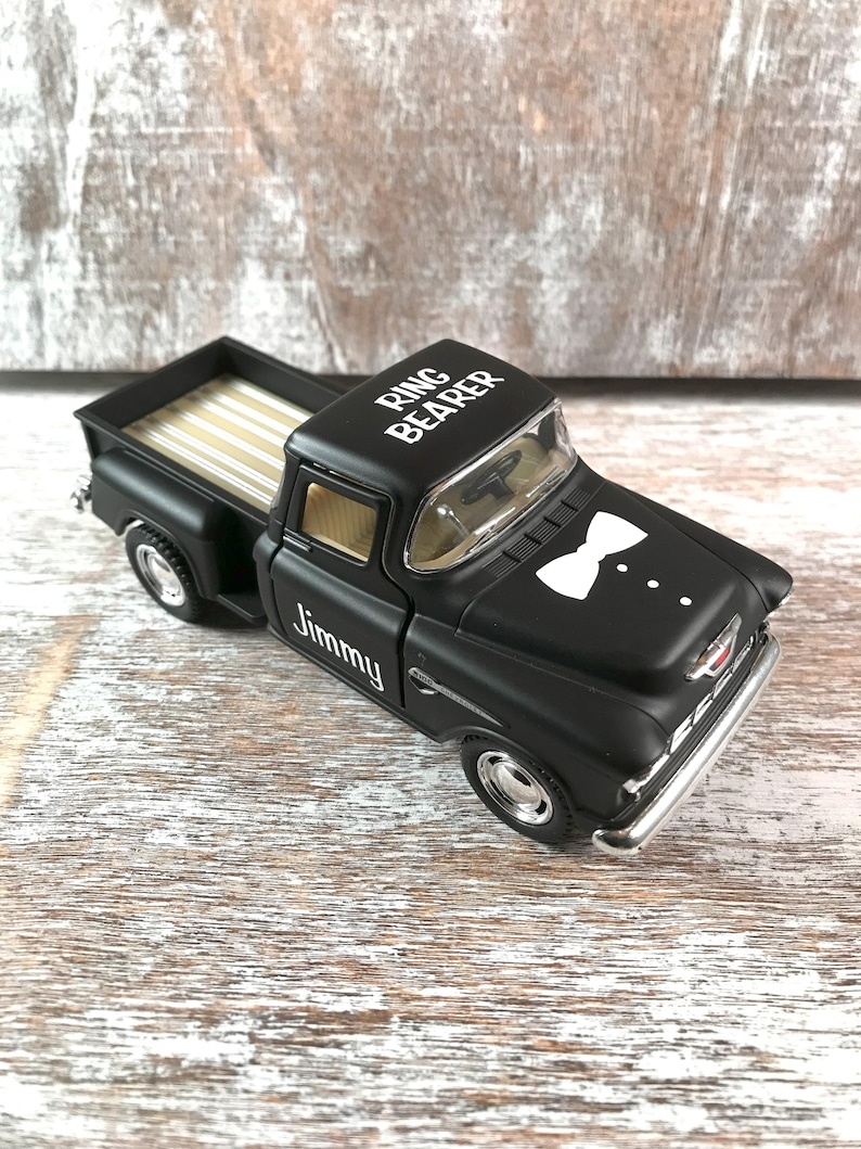 May include: A black toy truck with the words 'Ring Bearer' on the roof and 'Jimmy' on the side. The truck has a white bow tie design on the front. The truck is sitting on a light-colored wooden surface. The truck bed has a light-colored striped design.