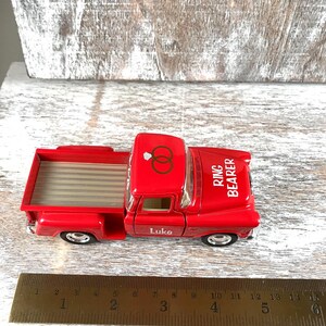 May include: A red toy pickup truck with the words "Ring Bearer" and "Luke" printed on it. The truck has a bed with a striped pattern and a roof with two interlocking rings. A ruler is next to the truck.