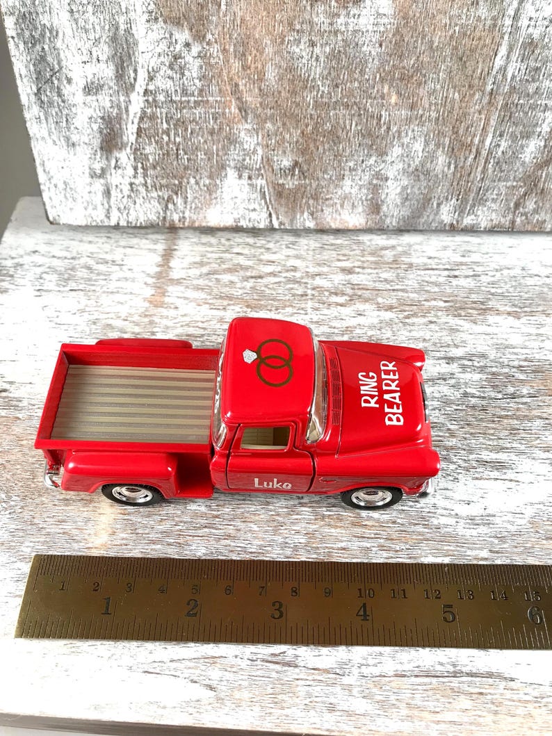 May include: A red toy pickup truck with the words "Ring Bearer" and "Luke" printed on it. The truck has a bed with a striped pattern and a roof with two interlocking rings. A ruler is next to the truck.