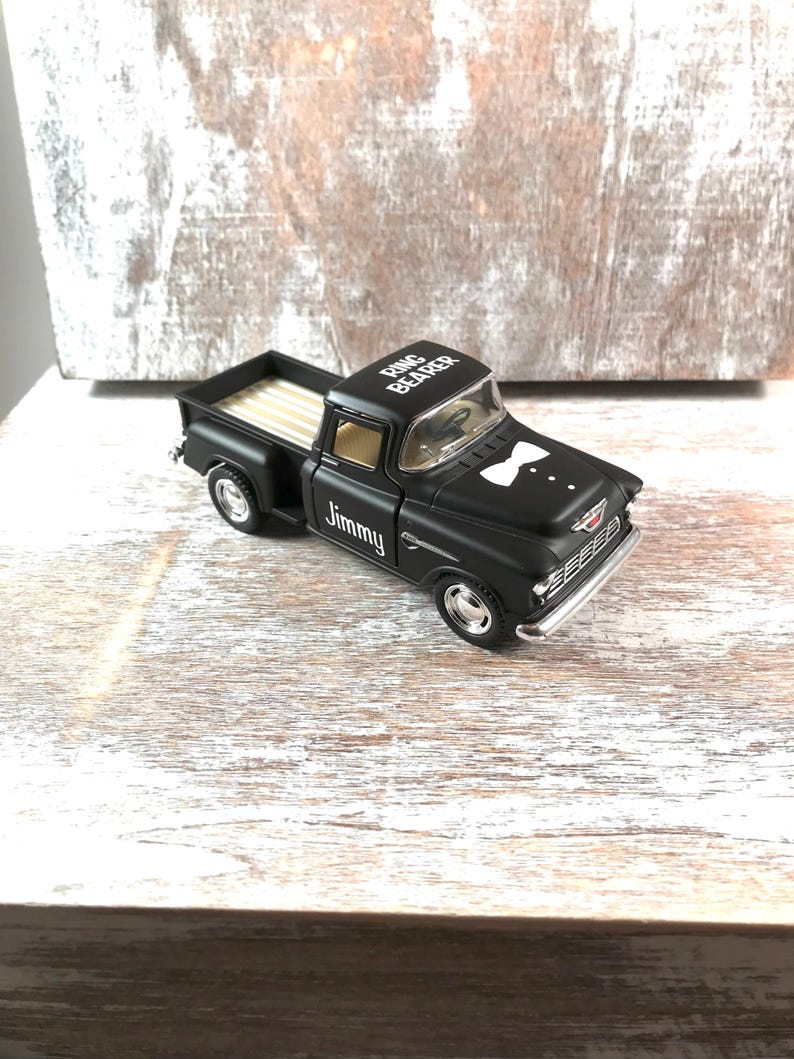 May include: A black toy pickup truck with the text "Ring Bearer" on the roof and "Jimmy" on the side. The truck has a white bow tie graphic and a tan bed. The truck is on a wooden surface.